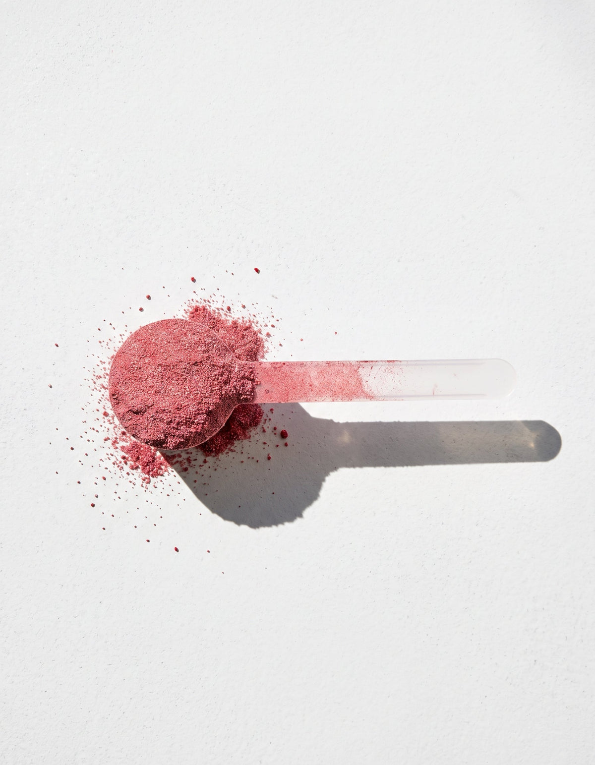 A clear plastic scoop filled with loose, finely ground dusty rose cosmetic powder, with some powder spilled around it, set against a clean white background with a sharp shadow.