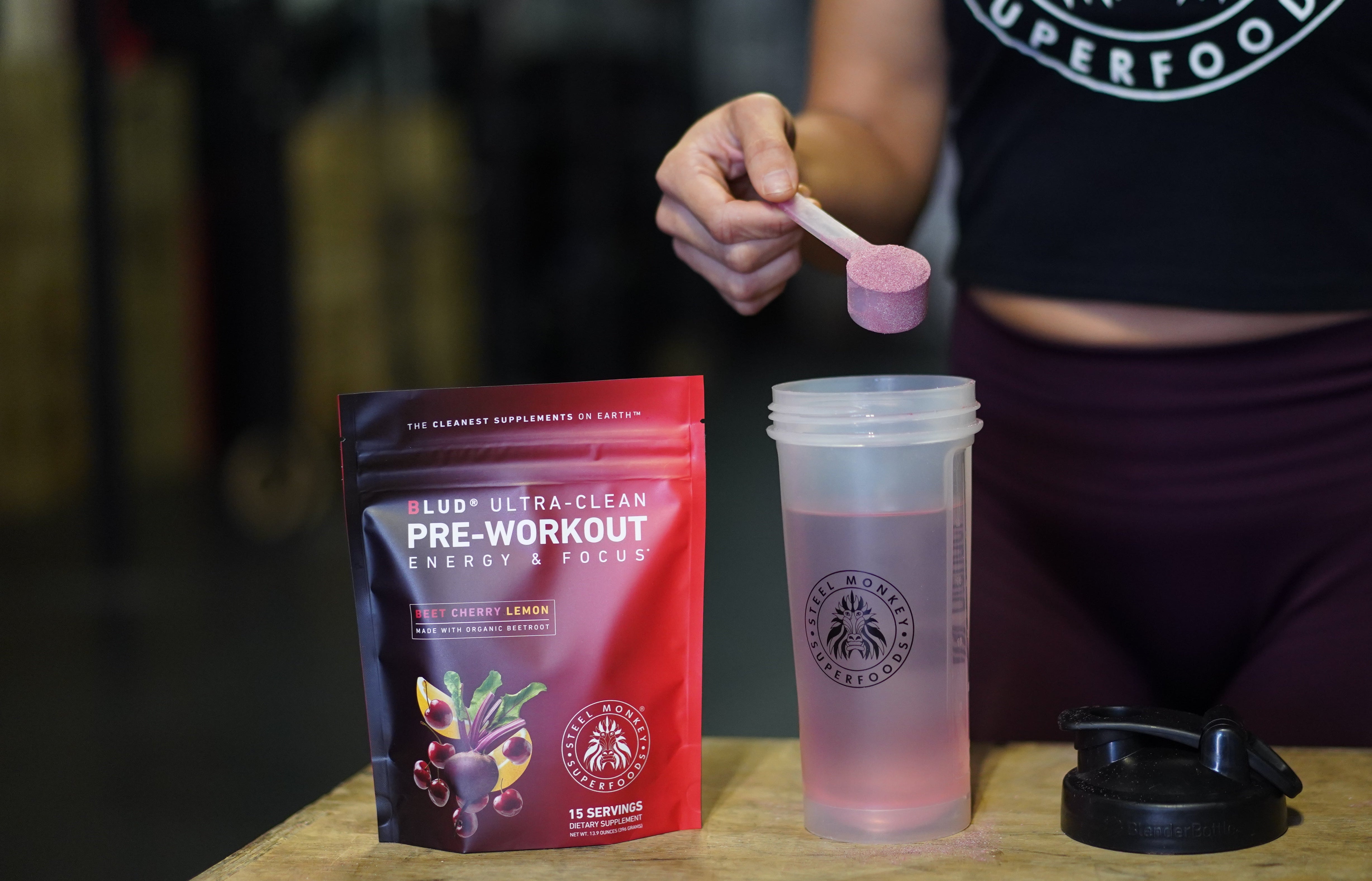 Person preparing a pre-workout supplement with a scoop and container on a table, featuring SuperFoods branding.