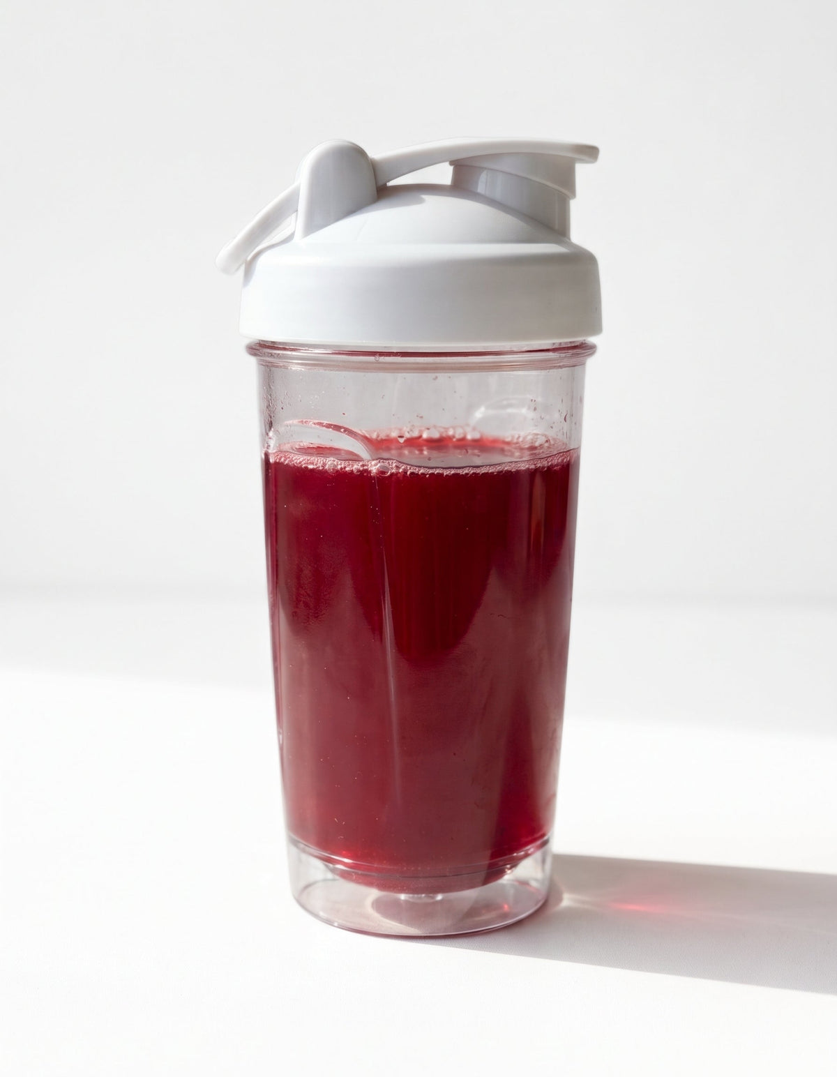A clear plastic shaker bottle with a white flip-top lid, filled with a deep red liquid, set against a bright white background with a soft shadow.