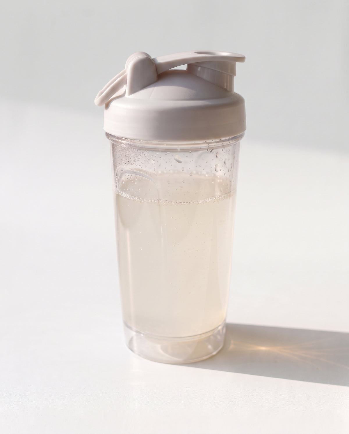 A clear plastic shaker bottle with a light beige lid and carrying loop, filled with a pale liquid, set against a bright white background with a soft shadow.